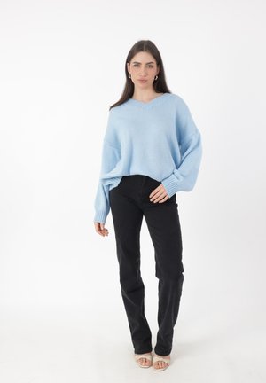 Lichtblauwe oversized trui met een v-hals, ribgebreide manchetten en een relaxed fit, gedragen met zwarte rechte jeans en open teen sandalen.
