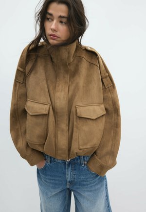 Frau trägt eine braune Wildleder-Bomberjacke mit großen Klappentaschen und blaue Jeans, steht mit den Händen in den Taschen vor einem einfarbigen Hintergrund.