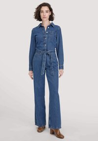 Frau in einem knöpfbaren Denim-Overall mit Taillengürtel, langen Ärmeln, Fronttasche und braunen Absatzstiefeln, vor einem schlichten Hintergrund stehend.