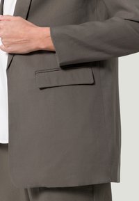 Blazer gris foncé avec une seule poche poitrine, texture lisse, structure ajustée et design épuré, associé à une chemise blanche en dessous.