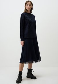 Maglione blu navy con ricamo floreale, abbinato a una gonna a pieghe blu navy e stivali militari neri con lacci, su uno sfondo neutro.