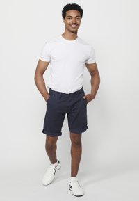 Jonge man die glimlacht, met zijn handen in zijn zakken, gekleed in een wit T-shirt, marineblauwe shorts en witte sneakers op een witte achtergrond.