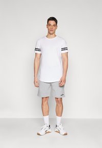 Camiseta blanca con rayas negras en las mangas, combinada con pantalones cortos grises claros. Zapatillas deportivas blancas con detalles en negro y marrón claro. Diseño sencillo.