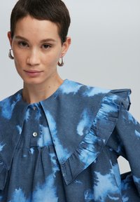 Blusa blu tie-dye con un grande colletto a volant, dettagli con bottoni e texture morbida. Presenta tonalità di blu variabili in un motivo a nuvola.