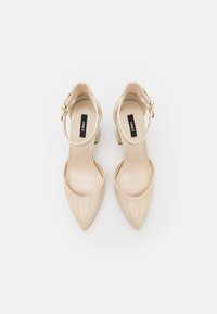 Beige kiilakuosiset korkokengät, joissa on terävä varvaskärki, nilkkaremmitys ja pieni lohkokorko. Kultaiset sisustuselementit remmin lähellä.