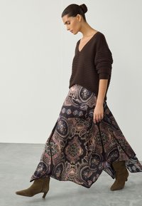 Pull en maille côtelée marron avec col en V, associé à une jupe maxi fluide à motifs dans des tons sombres et des bottes mi-hautes vert olive.