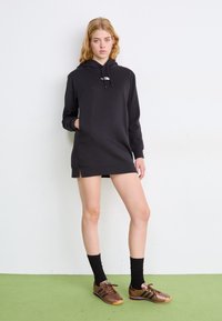 Μαύρο φόρεμα φούτερ με κουκούλα, μακριά μανίκια, μπροστινή τσέπη, πλαϊνές σχισμές και λεπτομέρεια με λογότυπο. Φορεμένο με καφέ αθλητικά παπούτσια και μαύρες κάλτσες.