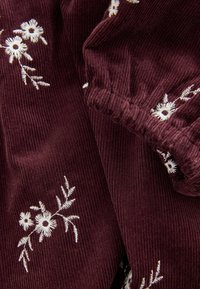 Burgunderfarbener Cordstoff mit weißer Blumenstickerei. Hat eine weiche Textur und elastische Ränder für eine geraffte Optik.
