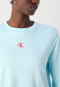 Hellblaues Crew-Neck-Sweatshirt aus weichem Stoff, mit einem rosa gestickten "ck"-Logo auf der Brust. Glatte Textur und lässige Passform.