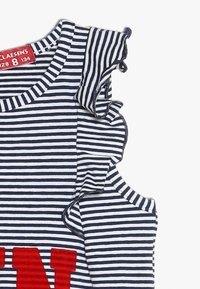 Randig skjorta med volangdetalj på axeln, med marinblå och vita ränder. Röd text på framsidan. Mjuk, strukturerad bomullstyg.