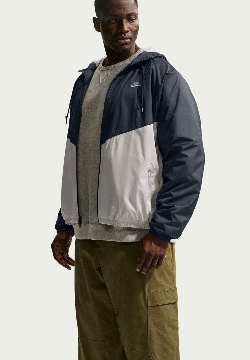 Man draagt een tweekleurige marineblauwe en witte Nike windjack, grijze trui en olijfgroene cargobroek, kijkt links naar beneden.