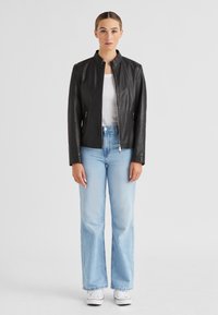 Schwarze Lederjacke mit Stehkragen, Reißverschluss und Seitentaschen, kombiniert mit hellblauen weit geschnittenen Jeans und weißen Sneakers.