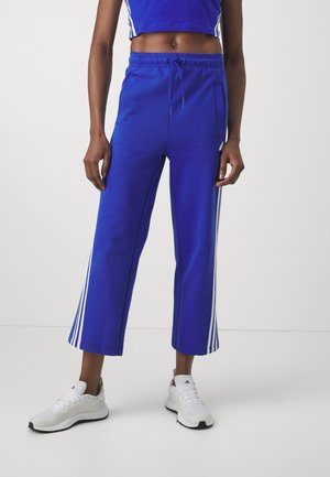 Persoon die blauwe cropped trainingsbroeken draagt met witte strepen aan de zijkanten, een bijpassende cropped top en witte sportschoenen op een witte achtergrond.