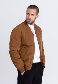 Bruine bomberjack met geribbelde manchetten en kraag, voorzien van een ritssluiting aan de voorkant en zijzakken, gedragen over een wit shirt. Gerenoveerde stof.