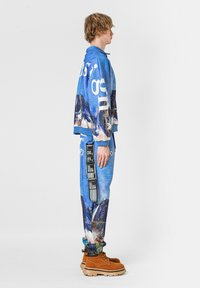 Sudadera gráfica azul con estampado de montaña y texto en blanco, combinada con joggers a juego que tienen un bolsillo lateral y una etiqueta negra. Botas marrones.