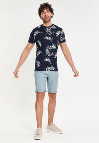 Camiseta de manga corta azul marino con patrón de hojas blancas, combinada con pantalones cortos azul claro enrollados y zapatillas gris claro. Textura de tela suave.
