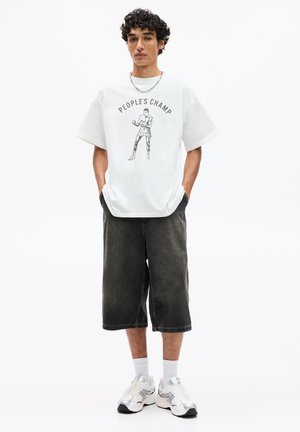 Tricou alb cu mânecă scurtă, cu imprimeu grafic "PEOPLE'S CHAMP" și figura, asortat cu pantaloni scurți din denim închis, până la genunchi, și adidași reflectorizanți.
