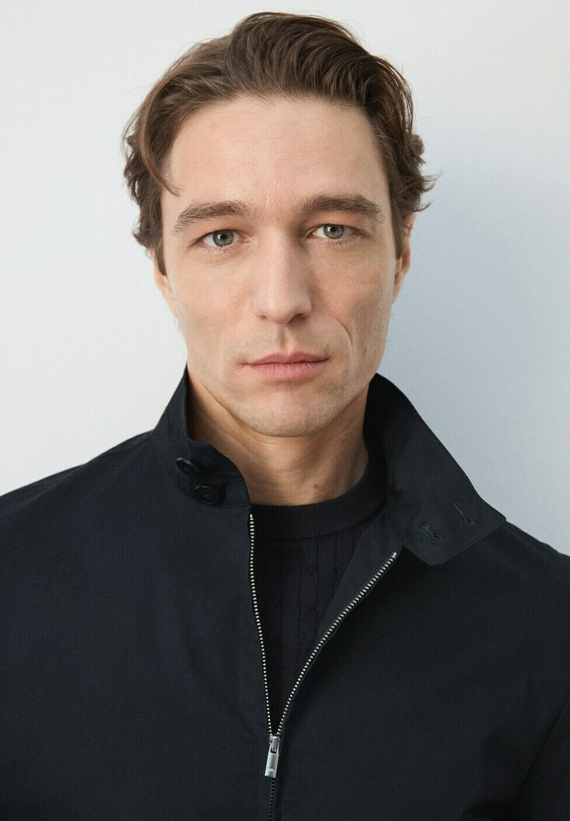 Homme à la peau claire et aux cheveux châtain clair, portant une veste noire zippée, debout devant un fond clair uni.