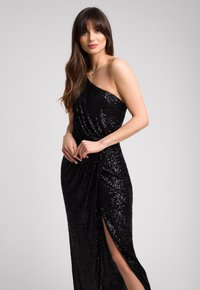 Kobieta z długimi brązowymi włosami, ubrana w czarną suknię cekinową na jedno ramię z wysokim rozcięciem, stojąca na tle jednolitego jasnego tła.