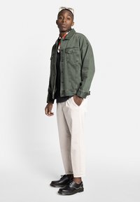 Olivenfarvet denimjakke, sort skjorte under, cremefarvede bukser og sorte sko med gule stikninger. Briller hviler på toppen af hovedet.