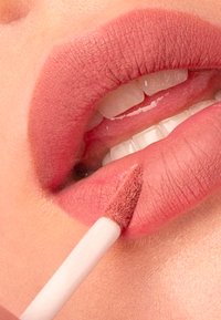 Flytande läppstift som appliceras på fylliga läppar. Färgen är en matt nude-rosa, med en mjuk textur och en noggrann applikatorspets synlig.