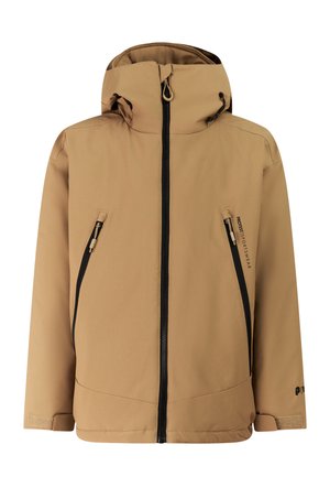 Jong persoon die een beige capuchonjas draagt, zwarte broek, zwarte muts en donkere schoenen, staand met handen in zakken tegen een eenvoudige lichte achtergrond.