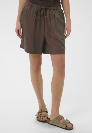 Vrouw die olijfgroene shorts met trekkoord en beige suède sandalen met dubbele gesp draagt, staand tegen een witte achtergrond.
