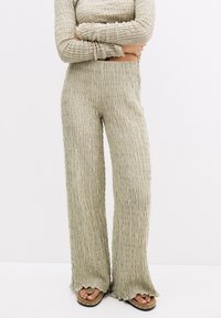 Beige gestructureerde wijde broek gecombineerd met een bijpassend lang mouwtop, model staat met gekruiste armen, blote voeten met studsandalen.