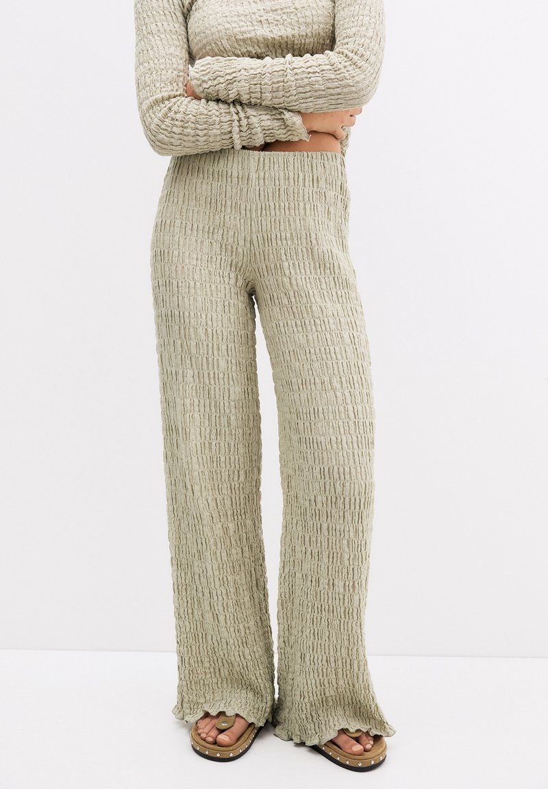 Beige gestructureerde wijde broek gecombineerd met een bijpassend lang mouwtop, model staat met gekruiste armen, blote voeten met studsandalen.