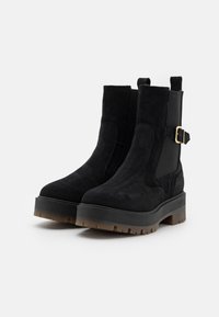 Bottines en suède noir avec des panneaux élastiques sur les côtés, accent de boucle en or, semelles épaisses en caoutchouc et profil chunky.