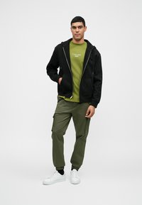 Μαύρο φούτερ με φερμουάρ πάνω από πράσινο T-shirt με λογότυπο, συνδυασμένο με ελιά cargo παντελόνι και λευκά αθλητικά παπούτσια, αναδεικνύοντας μια χαλαρή, στρωτή εμφάνιση.