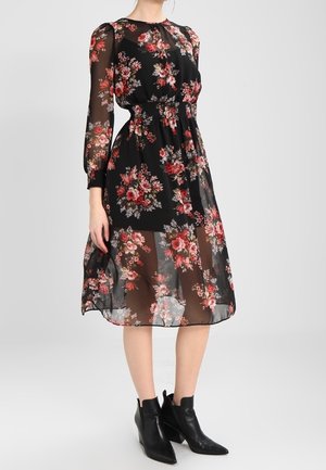 Frau trägt ein schwarzes, durchsichtiges Midikleid mit rotem und pinkem Blumenmuster und schwarzen Stiefeletten, steht vor einem einfarbigen grauen Hintergrund.