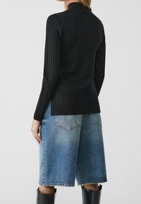 Persoon in een zwarte ribbelige lange mouwen top, losse blauwe denim korte broek tot de knie en zwarte laarzen tot de knie, staande tegen een effen achtergrond.