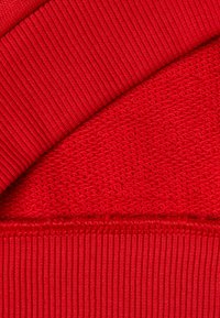 Červená pletenina s dvěma texturami: hladká, těsná žebrovaná část a volná, otevřená tkanina s pravidelným vzorem.