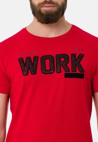 Rotes Baumwoll-T-Shirt mit auffällig schwarzem "WORK HARD"-Druck in strukturierter Ausführung. Kurze Ärmel und Rundhalsausschnitt.