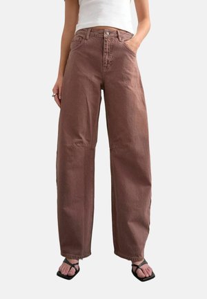 Persoon draagt bruine wijde broek met hoge taille en zakken, wit mouwloos topje en zwarte sandalen met bandjes en vierkante neus tegen een effen achtergrond.