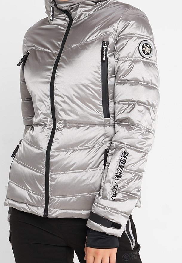 Silvrig puffer-jacka med en figursydd silhuett, fickor med dragkedja och svarta detaljer. Inkluderar logotyp på ärmen och strukturerad tyg.