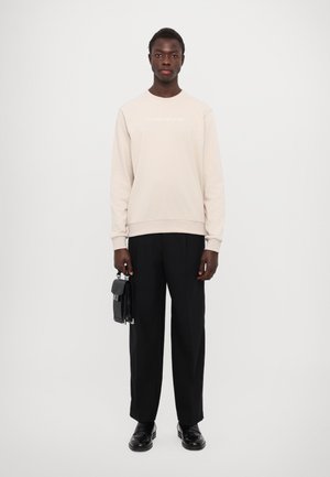 Beige sweatshirt met ronde hals, geborduurde tekst, gecombineerd met zwarte broek en een kleine zwarte tas, staande voor een neutrale achtergrond.