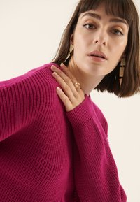 Pull en maille fuchsia avec texture côtelée et manches longues. Accessoires dorés et gros plan sur une main portant une bague.