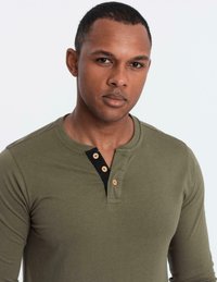 Man med kort hår som bär en olivgrön långärmad henley-tröja med tre träknappar, som tittar in i kameran mot en vit bakgrund.