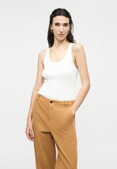 Γυναίκα με σκούρα μαλλιά που φοράει χρυσούς κρίκους στα αυτιά, λευκό αμάνικο μπλουζάκι και καφέ παντελόνι, στέκεται με το ένα χέρι στην τσέπη μπροστά σε φωτεινό φόντο.