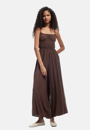 Brown, sleeveless jumpsuit with a smocked bodice and wide-legged design, made from lightweight fabric. Worn with flat, light-colored shoes.