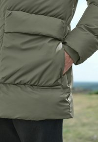 Olivinės spalvos paminkštinta striukė su didele priekinėmis kišenėmis ir guminėmis rankovėmis, pagaminta iš lygios, blizgios medžiagos su horizontaliomis siūlėmis.