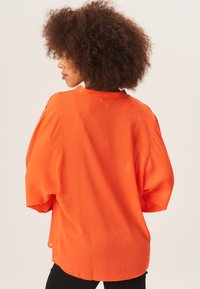 Oversized orange blouse with pleated texture, rounded neckline, and wide sleeves. The fabric appears lightweight and semi-sheer.
