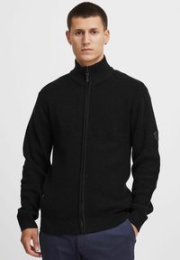 Suéter negro de punto con cremallera y cuello alto, de tejido texturizado, con puños y bajo acanalados, que presenta un parche con logo en la manga.