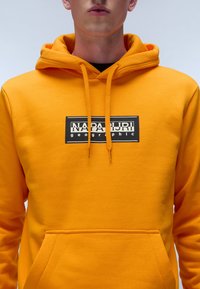 Geel hoodie van zacht materiaal met een frontzak, trekkoord capuchon en een zwart logo in een rechthoekig ontwerp.