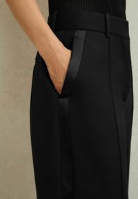 Roka, kas atpūšas iekšā priekšējā kabatā melniem pielāgotiem biksēm ar satīna apmalēm kabatu malās un jostasvietā.