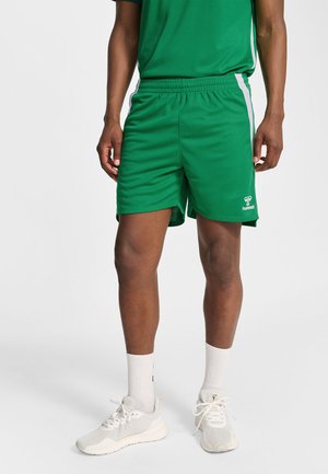 Mand iført grønne atletikshorts med hvide sidelister, hvide strømper og grå løbesko, stående foran en ensartet baggrund.