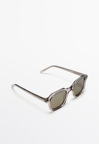 Okulary przeciwsłoneczne z okrągłymi oprawkami wykonanymi z szarego, półprzezroczystego tworzywa sztucznego, ciemnymi soczewkami oraz smukłymi metalowymi zdobieniami na zausznikach.