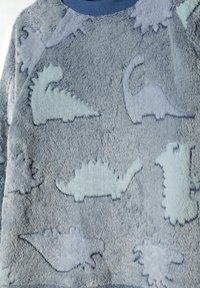 Sweatshirt gris pelucheux arborant des silhouettes de dinosaures bleus variés, avec une texture douce et des accents bleu clair sur un fond atténué.